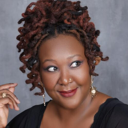 A headshot of Saphira Joy. She has a fun nest of curly red locs in an updo, warm red lipstick, a bright smile, and is looking up and to the left.